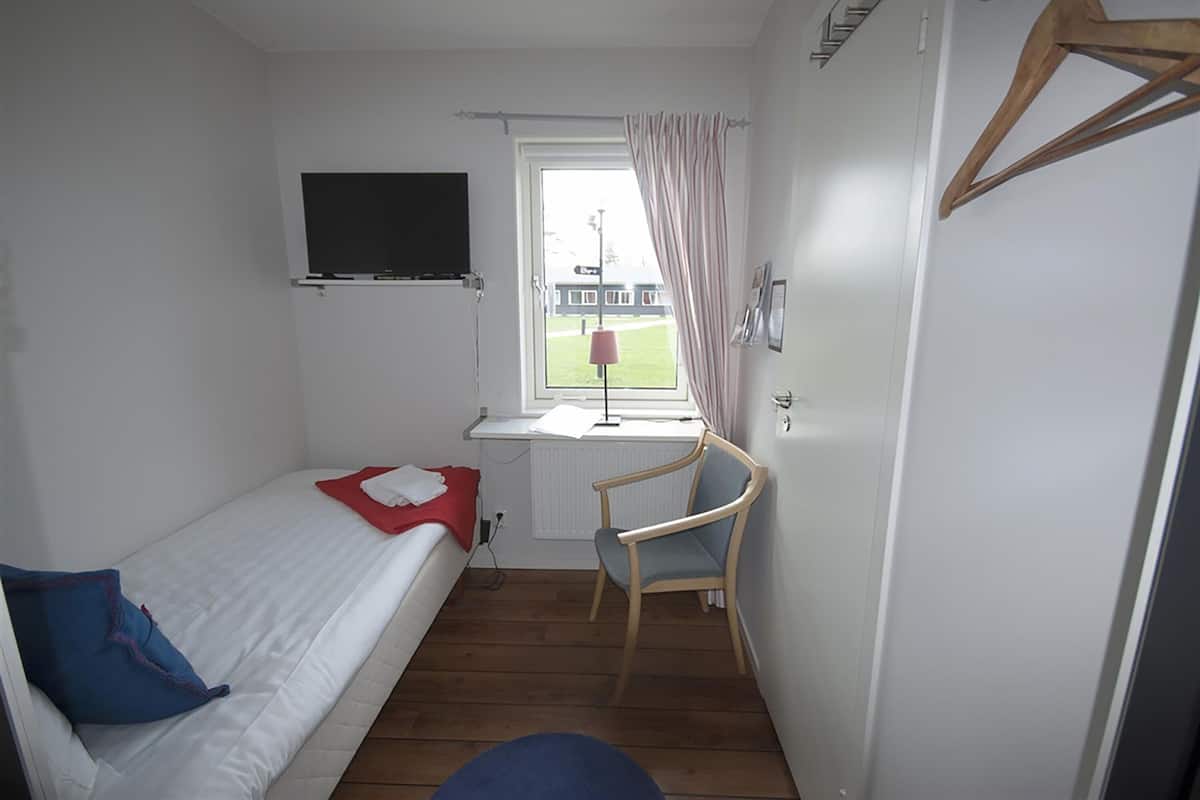 A small, well-lit room with a single bed, a desk with a chair, a television mounted on the wall, and a window with curtains.