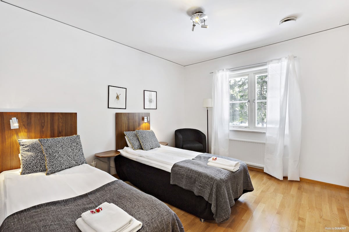 A hotel room with two beds, a wooden headboard, a window with white curtains, and a small table with a lamp.