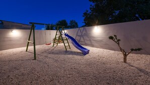 Une aire de jeux avec un toboggan et des balançoires, entourée d’un mur et d’un petit arbre.