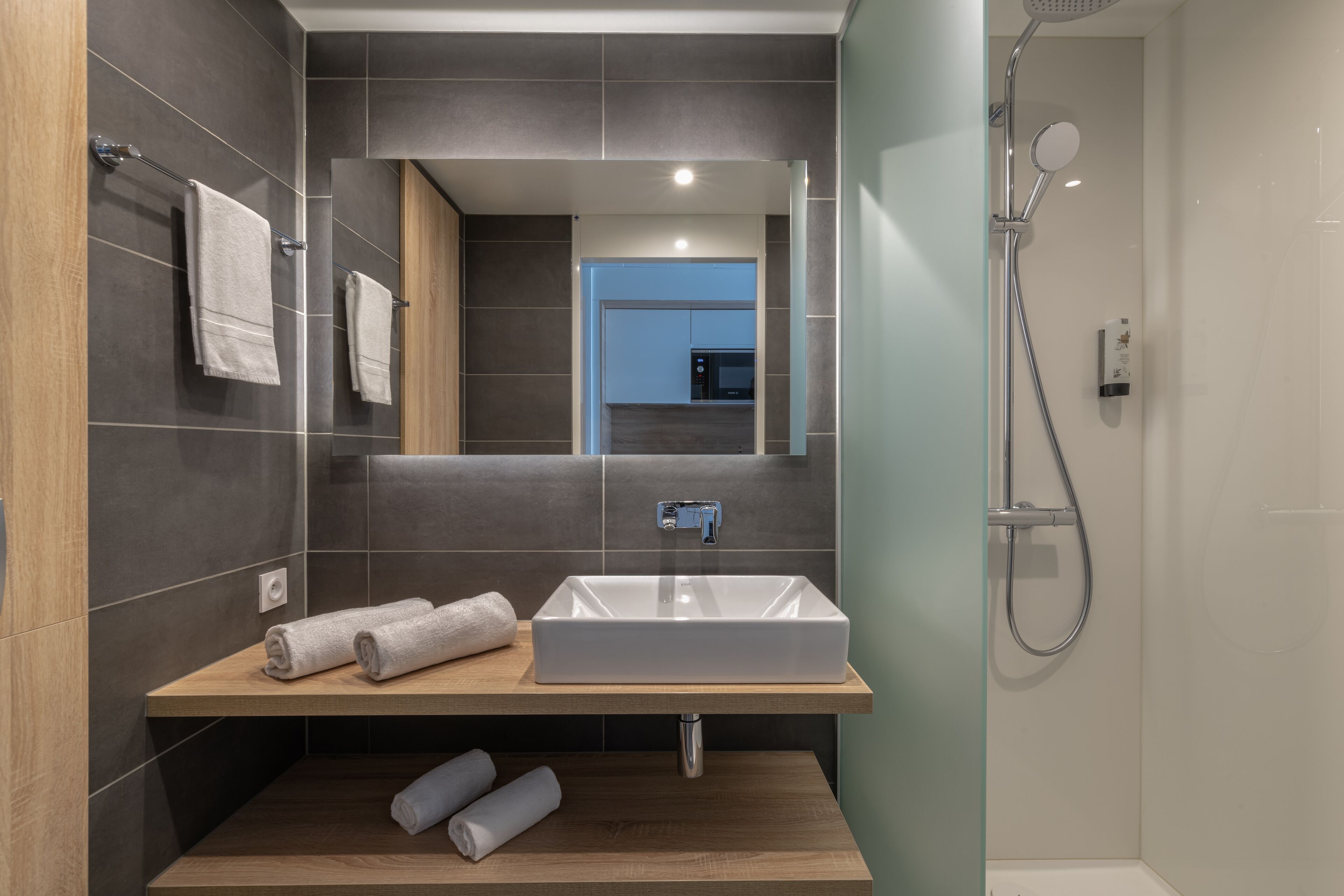 A modern bathroom with a rectangular sink, wooden vanity, dark tiled walls, a shower with a handheld showerhead, and a visible kitchen area in the background.