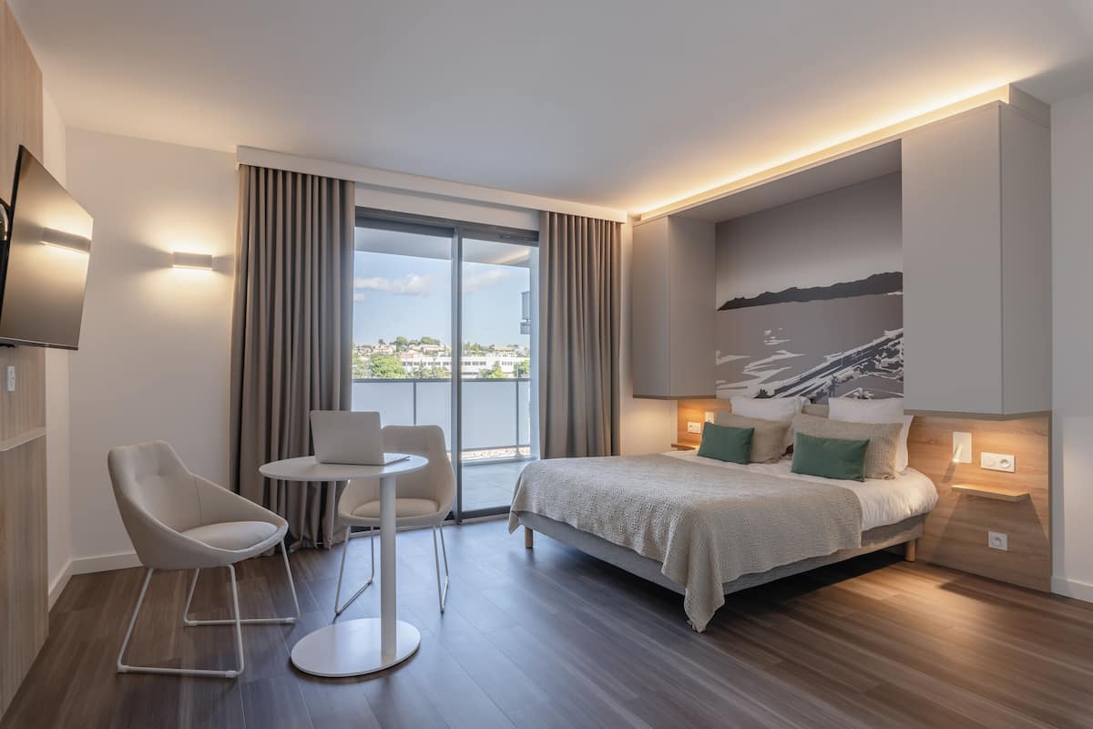 Ein modernes Hotelzimmer mit einem Bett, zwei Sesseln, einem kleinen Tisch und einem großen Fenster mit Vorhängen.