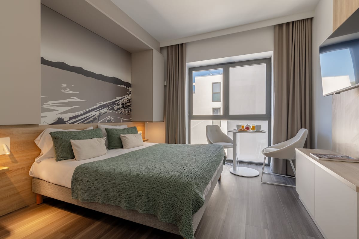 Ein modernes Hotelzimmer mit einem großen Bett, einem Schreibtisch und einem Sessel. Ein Fenster mit Vorhängen, ein Bild an der Wand und ein Blick ins Freie.