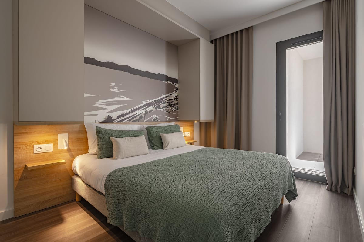 Ein modernes Hotelzimmer mit einem großen Bett, einem Nachttisch, einer Wandleuchte und Blick ins Freie durch ein Fenster.