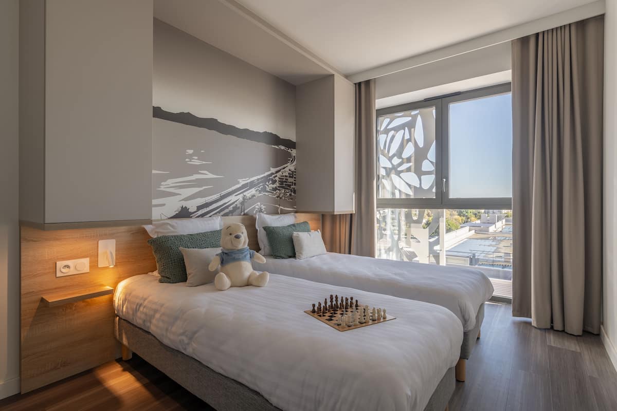 Ein modernes Hotelzimmer mit zwei Betten, einem großen Fenster mit Vorhängen, einem Wandbild und einem Schachbrett auf dem Bett.