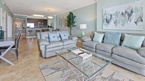 A modern living room with two grey sofas, a glass coffee table, and a dining area with a white table and chairs. A kitchen is visible in the background with a bar counter and stools. - Gorgeous 11th Floor, Corner Unit, 4 Bdrm Condo, Two Sets of FREE Beach Chairs (Panama City Beach)