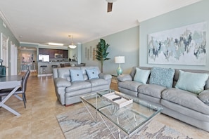 A modern living room with two grey sofas, a glass coffee table, and a dining area with a white table and chairs. A kitchen is visible in the background with a bar counter and stools.