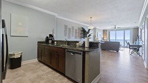 A modern kitchen with a dishwasher, dark wooden cabinets, and a marble countertop. There is a dining area with a sofa and a table in the background. - Gorgeous 11th Floor, Corner Unit, 4 Bdrm Condo, Two Sets of FREE Beach Chairs (Panama City Beach)