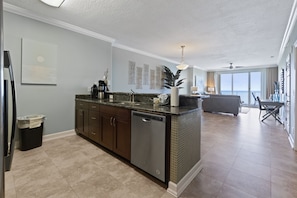A modern kitchen with a dishwasher, dark wooden cabinets, and a marble countertop. There is a dining area with a sofa and a table in the background.