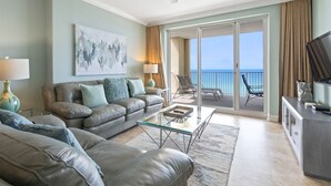 A spacious living room with a sofa, a glass coffee table, and a television on a cabinet. A large painting adorns the wall, and a balcony with a view of the ocean is visible. - Gorgeous 11th Floor, Corner Unit, 4 Bdrm Condo, Two Sets of FREE Beach Chairs (Panama City Beach)