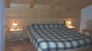 Een slaapkamer met een houten plafond, een bed met een geruit plaid, twee nachtkastjes met lampen, en een stoel.
