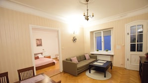 A room with a wooden herringbone floor, a sofa, a dining table with chairs, a window with a view, and a chandelier. - Elegant Apartment Universitas (Warsaw)