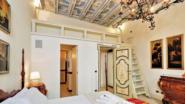 A hotel room with a large bed, a wooden dresser, a chandelier, and a staircase leading to an upper level.