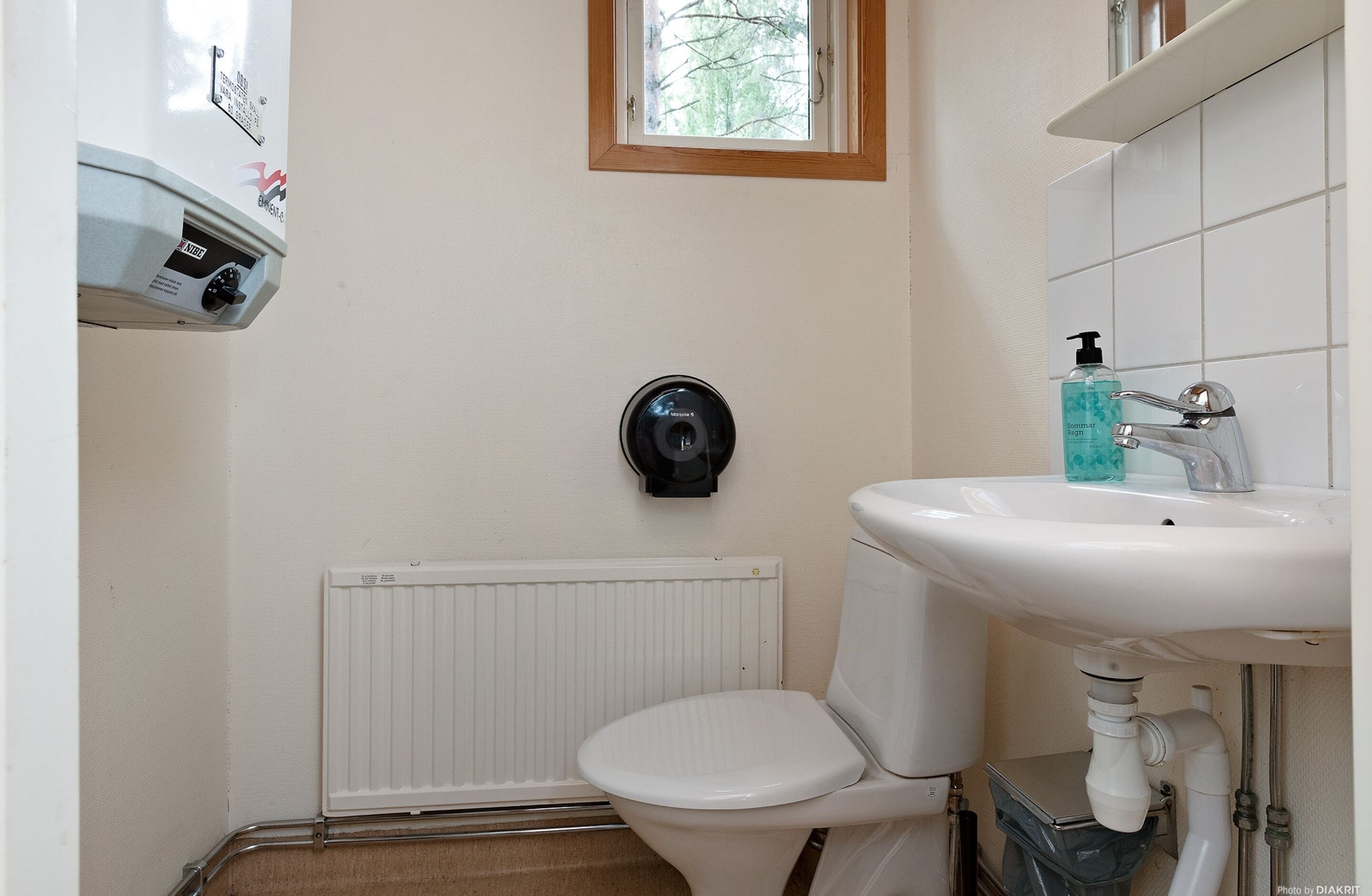 Un baño pequeño con inodoro, lavamanos blanco, secamanos y una ventana sobre el inodoro.