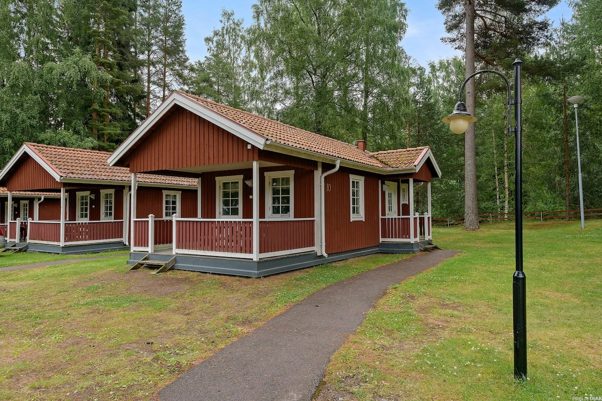 Une rangée de cabanes en bois, dotées d’un porche et numérotées de 1 à 10, est aménagée dans une zone boisée, et un chemin pavé y mène.