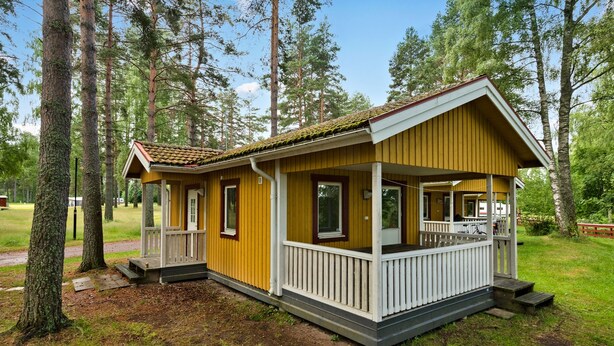 Une cabane jaune avec un porche, entourée d’arbres.