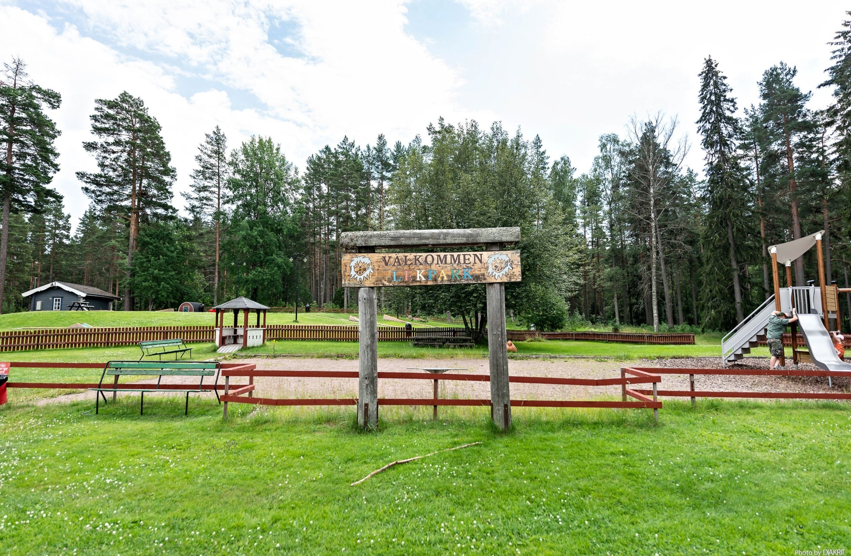 A park with a wooden sign, picnic tables, a playground, and a fenced area.
