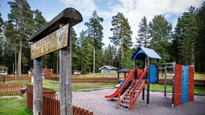 Une aire de jeux avec un toboggan et des structures d’escalade, entourée d’une clôture en bois et d’un panneau indiquant : « Valkommen Lkpark ».