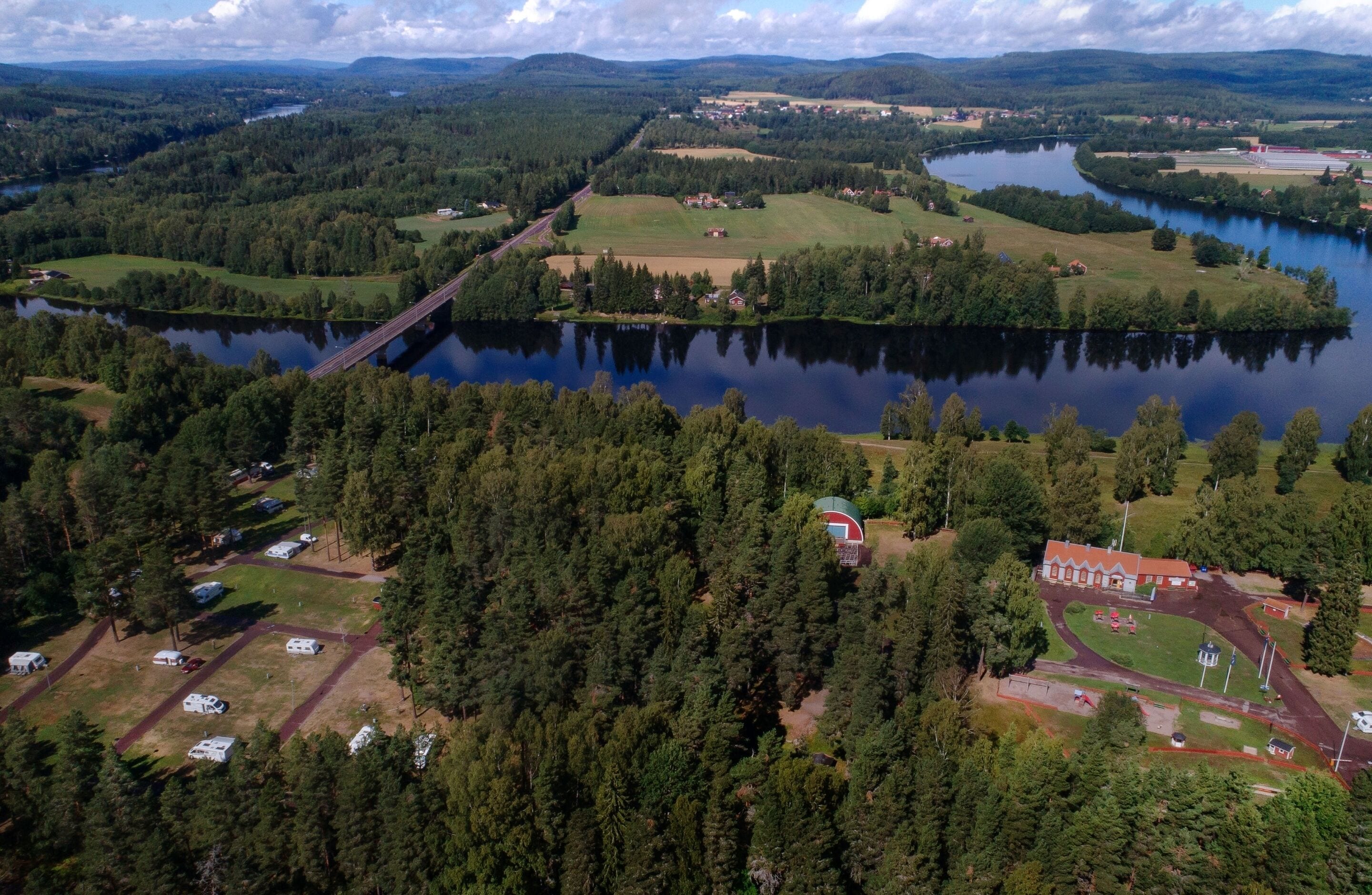 Ett flygbildsbild av ett område vid en sjö med en bro, skogsområden och en liten grupp av byggnader.