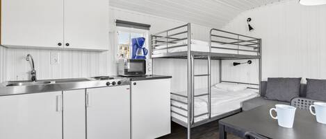 A compact kitchen with a sink, stove, and microwave. There is a bunk bed with a table and chairs in the room.