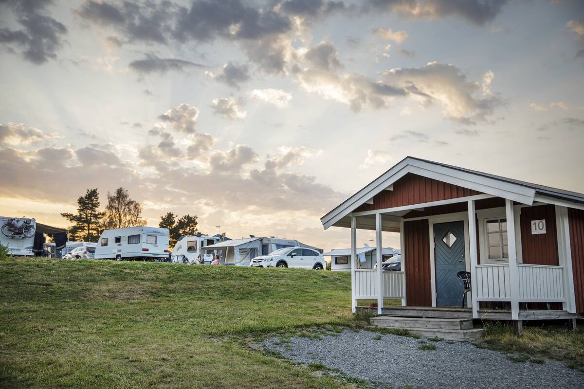 Eine kleine Holzhütte mit Veranda, Nummer 10, umgeben von Wohnmobilen und Autos auf einem Campingplatz.