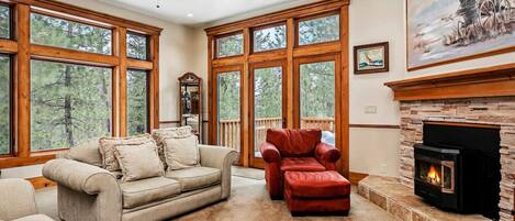 A spacious living room with large windows, a fireplace, a ceiling fan, and a painting on the wall.