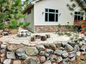 Relax on the patio surrounded by a serene garden and pine trees, perfect for outdoor dining or unwinding.