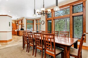 Enjoy meals in this dining area with a large table and six chairs, surrounded by a high ceiling and large windows offering a view of the surrounding forest.
