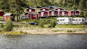 Et område med hytter ved søen, hvor der er røde hytter, en hvid campingvogn og en sort bil parkeret tæt på vandet.