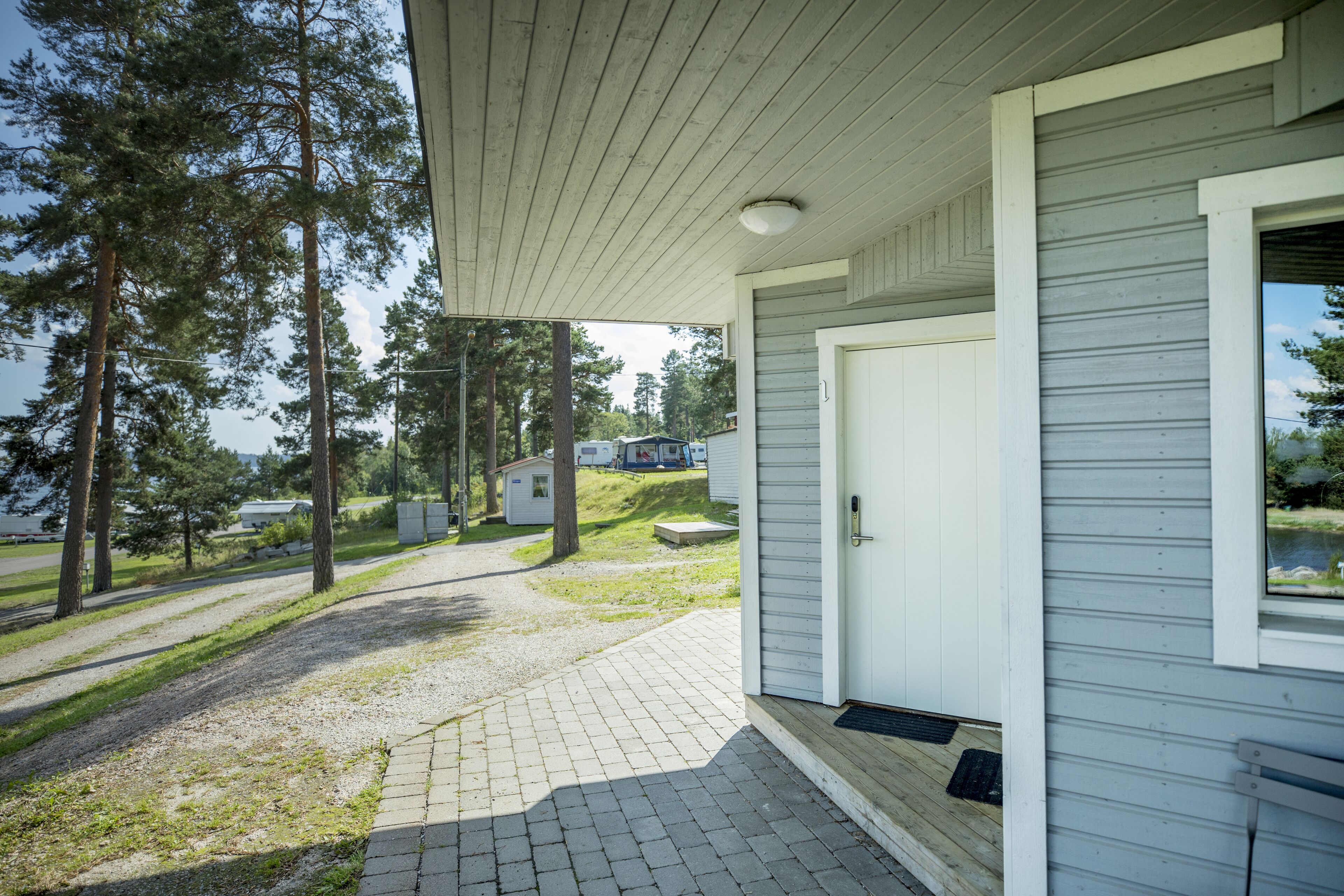 A covered porch with a wooden ceiling, a white door, and a view of a grassy area with trees and a small building in the distance.