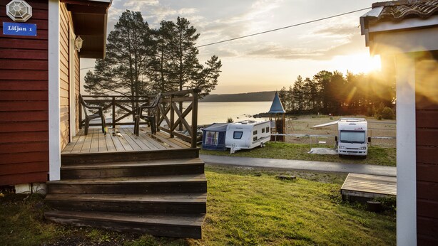 En træhytte med veranda og trapper til en terrasse, en hvid autocamper parkeret i nærheden, samt et klart udsyn til vand og træer i baggrunden.
