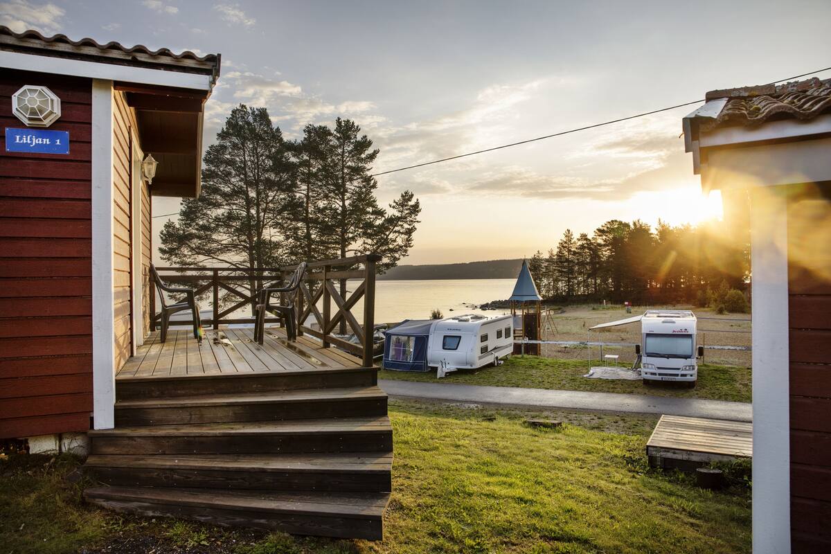 Een houten blokhut met een veranda en treden die leiden naar een terras, een witte caravan in de buurt, en een helder uitzicht op het water en de bomen op de achtergrond.