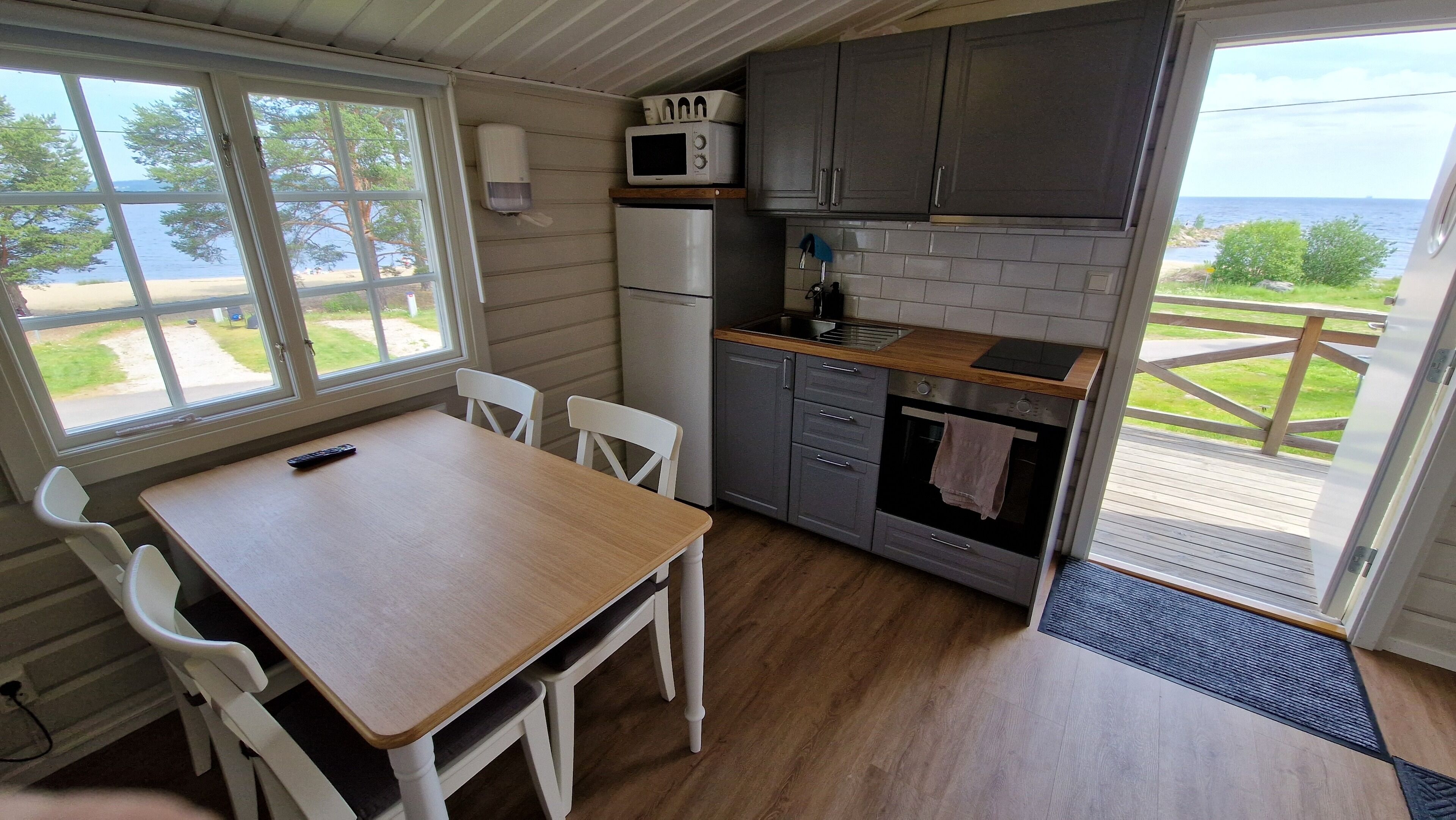 A compact kitchen with a wooden table and chairs, a microwave, a refrigerator, and a view of the sea through a large window.