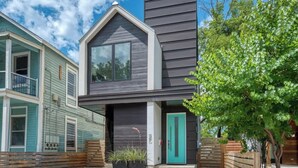 Une maison moderne à deux étages, avec une façade sombre, une porte de couleur turquoise et un petit jardin devant, agrémenté d’un arbre et de plantes.