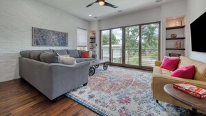 Un salon moderne avec un canapé gris, un canapé beige et une table basse. Une grande fenêtre offre une vue sur les arbres, et un ventilateur de plafond est installé au centre de la pièce.