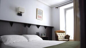 Une chambre d’hôtel avec un grand lit, une table de chevet avec une lampe, un tableau encadré au mur et une chaise près de la fenêtre.