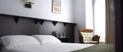 A hotel room with a large bed, a bedside table with a lamp, a framed picture on the wall, and a chair by the window.