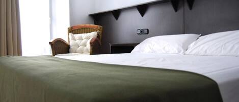 A hotel room with a large bed, a side table, a chair, and a headboard with a control panel.