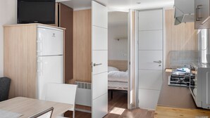 Una habitación compacta con cama, una kitchenette pequeña, un televisor encima de un mueble, y una puerta que da a otra habitación.