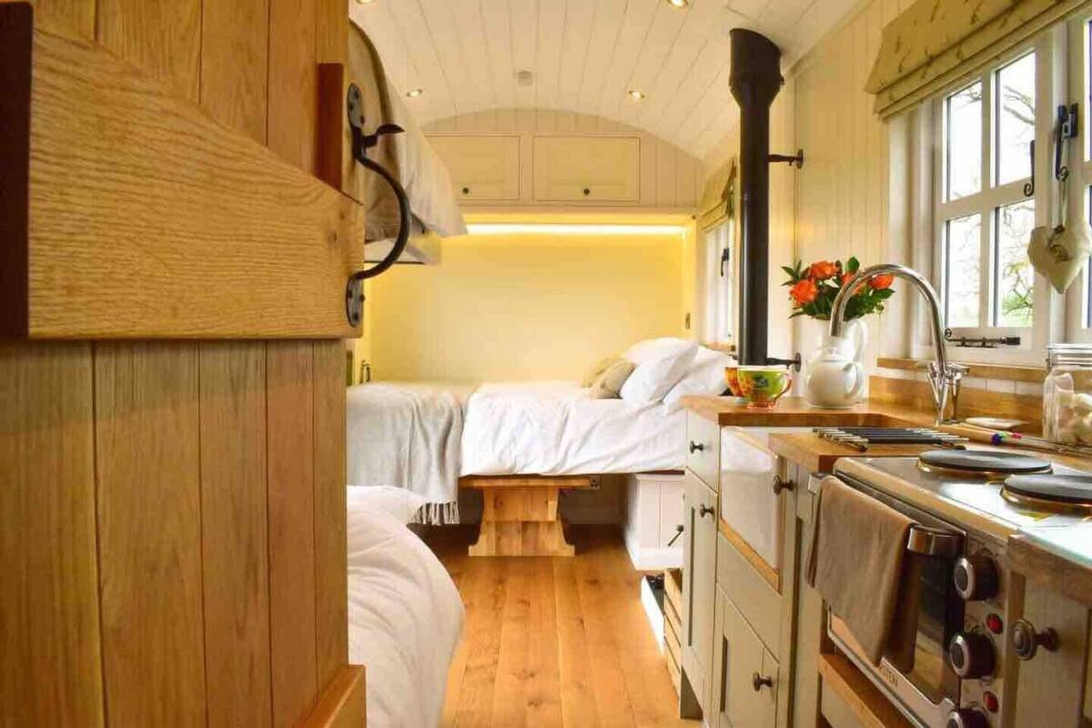 A cozy cabin interior with wooden walls, a bed, a small table, and a kitchen area with a stove and sink.