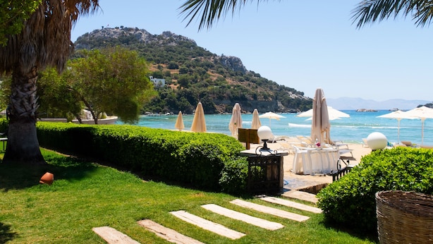Um alpendre à beira-mar com guarda-sóis, uma mesa e cadeiras, com vista para o mar azul e um terreno acidentado.