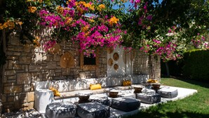 Área externa com assentos, paredes de pedra, almofadas e um jardim com flores rosa e laranja.