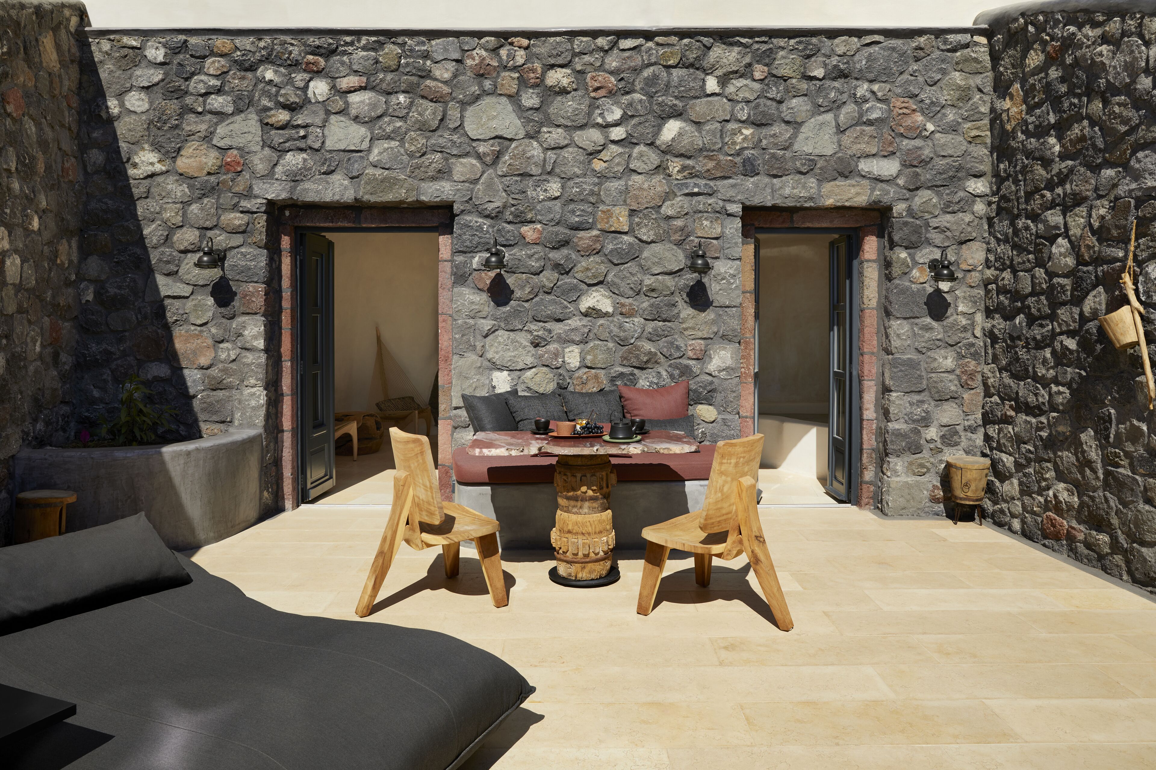 A stone-walled outdoor seating area with two wooden chairs, a small table, and a sofa.