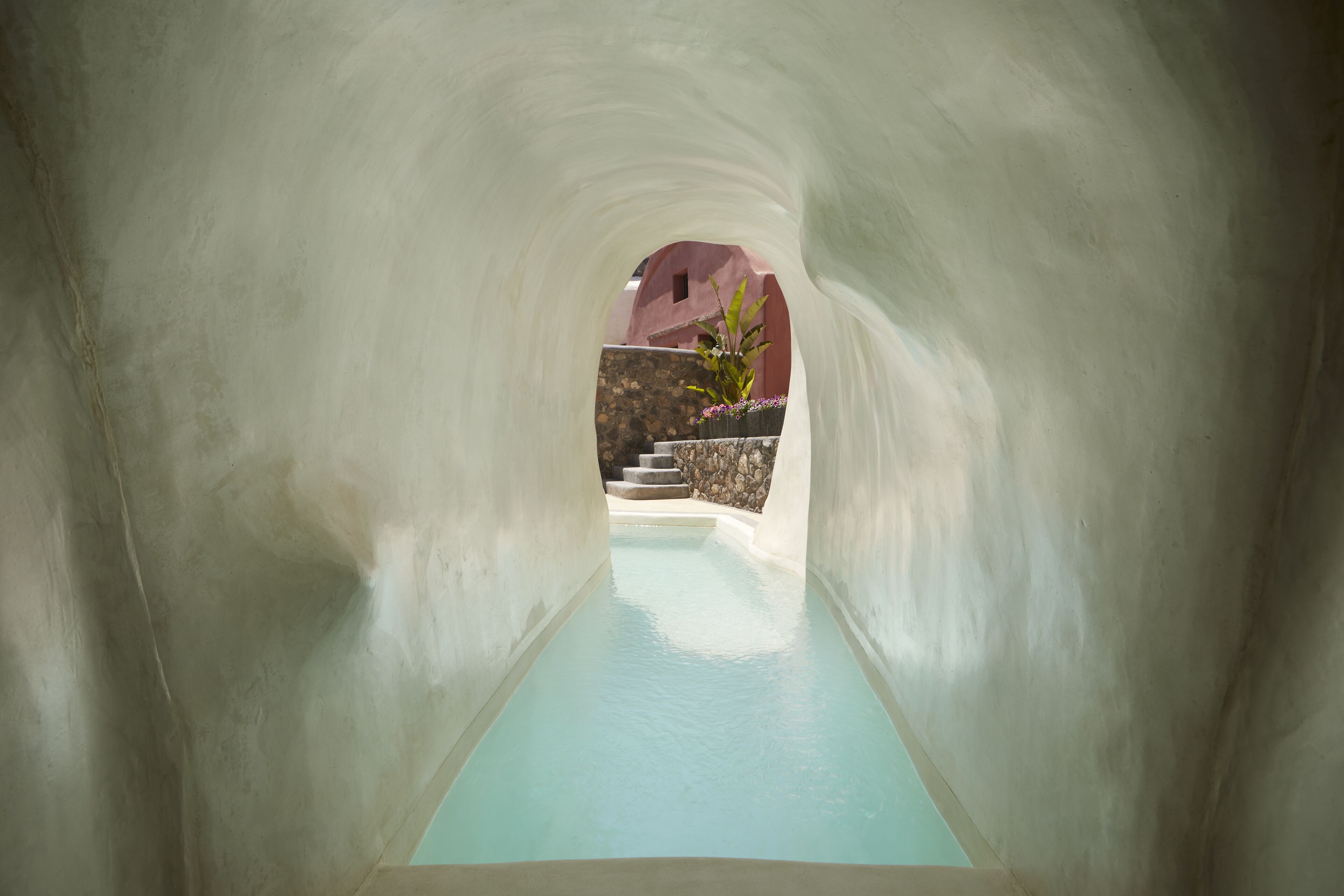 A tunnel leading to a pool with a stone wall and stairs in the background.