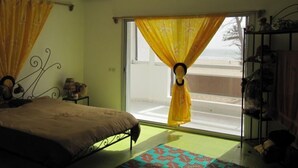 A bedroom with a bed, a window with yellow curtains, a small table with a lamp, and a shelf with items. - Eden Beach Saint-Louis (St. Louis)