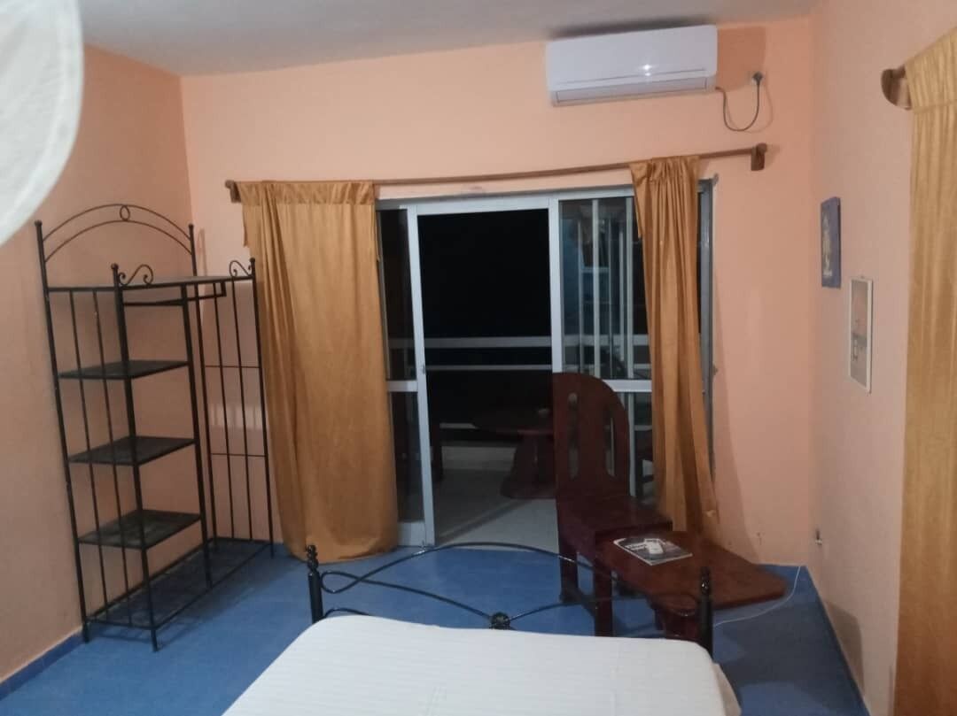 A hotel room with a bed, a metal shelf unit, a chair, and a sliding glass door leading to a balcony.