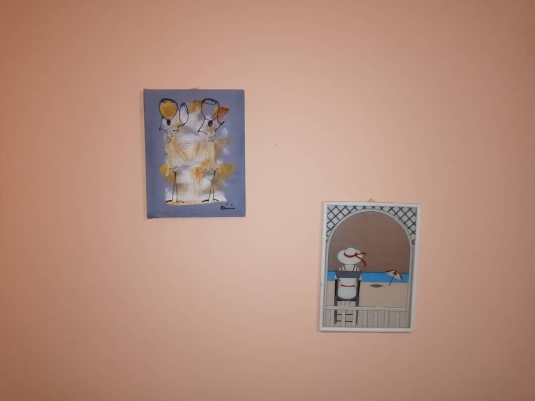 Deux œuvres d’art fixées au mur : l’une est une composition abstraite aux formes jaunes et blanches, et l’autre représente une silhouette vêtue de blanc et coiffée d’un chapeau rouge, se tenant devant un fond bleu.