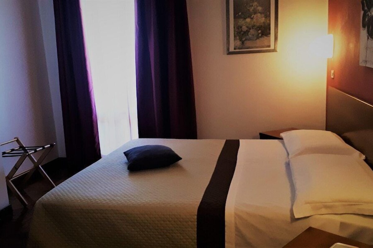 Ein Hotelzimmer mit einem Bett, einem Nachttisch, einer Lampe, einem Gemälde an der Wand und einem Fenster mit Vorhängen.