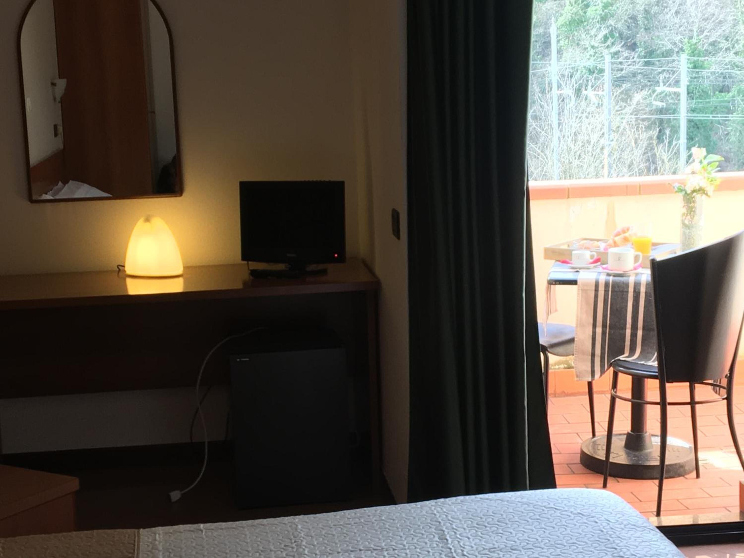 Quarto de hotel com cama, escrivaninha com luminária, televisão e vista para o exterior pela janela.