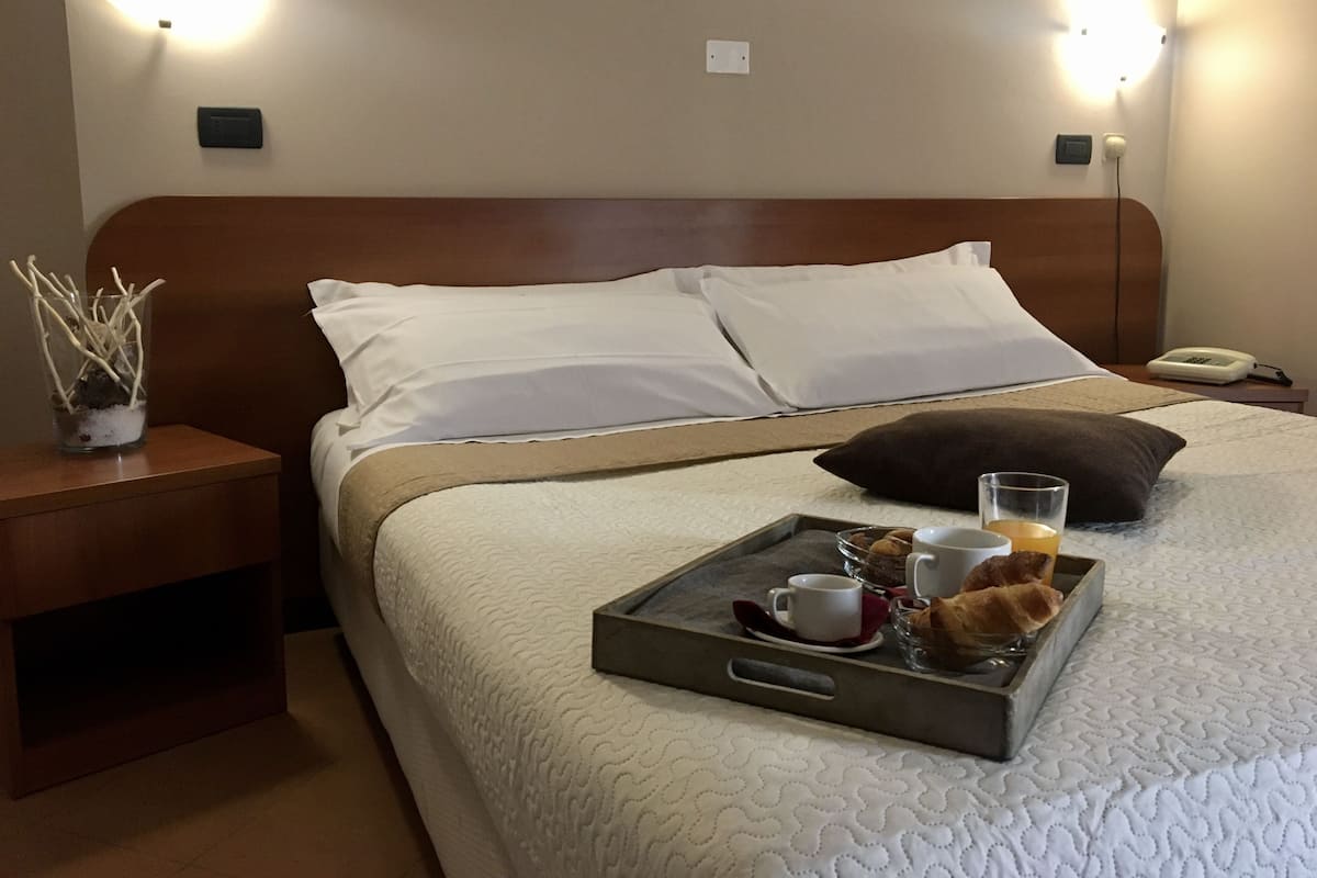 Un lit bien fait, avec du linge de lit blanc, une assiette avec des croissants, une tasse et un verre de jus d’orange posés sur le lit.