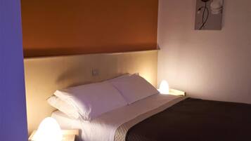 Una cama bien hecha con sábanas blancas y una manta oscura, una lámpara de noche y una lámpara de pared.
